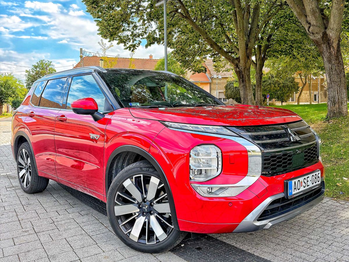 Piros Mitsubishi Outlander PHEV plug-in hibrid SUV parkol egy fákkal szegélyezett utcán.