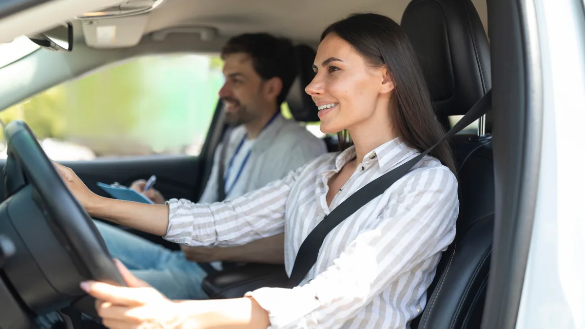 Cheerful confident pretty young woman sitting inside car with man instructor by her side, lady student holding hands on steering wheel, passing exams successfully at driving school, getting licence
