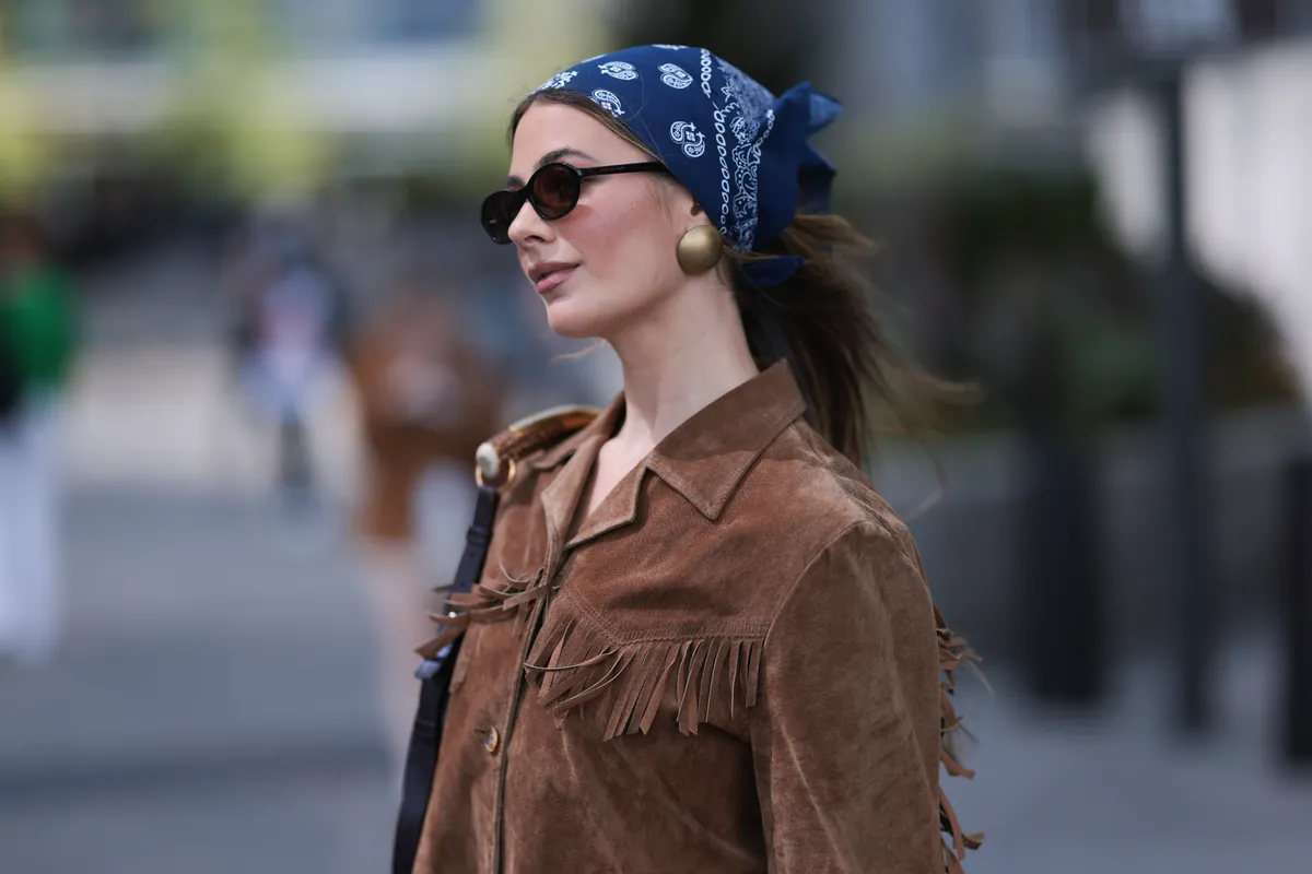 NEW YORK, NEW YORK - SEPTEMBER 08: A guest seen wearing a blue pattern bandana, gold round earrings, DMY Studios black oval sunglasses, vintage brown suede leather jacket with fringe details and brown leather shoulder bag, before the Jason Wu Collection show, during the New York Fashion Week, on September 08, 2024 in New York City. (Photo by Jeremy Moeller/Getty Images)