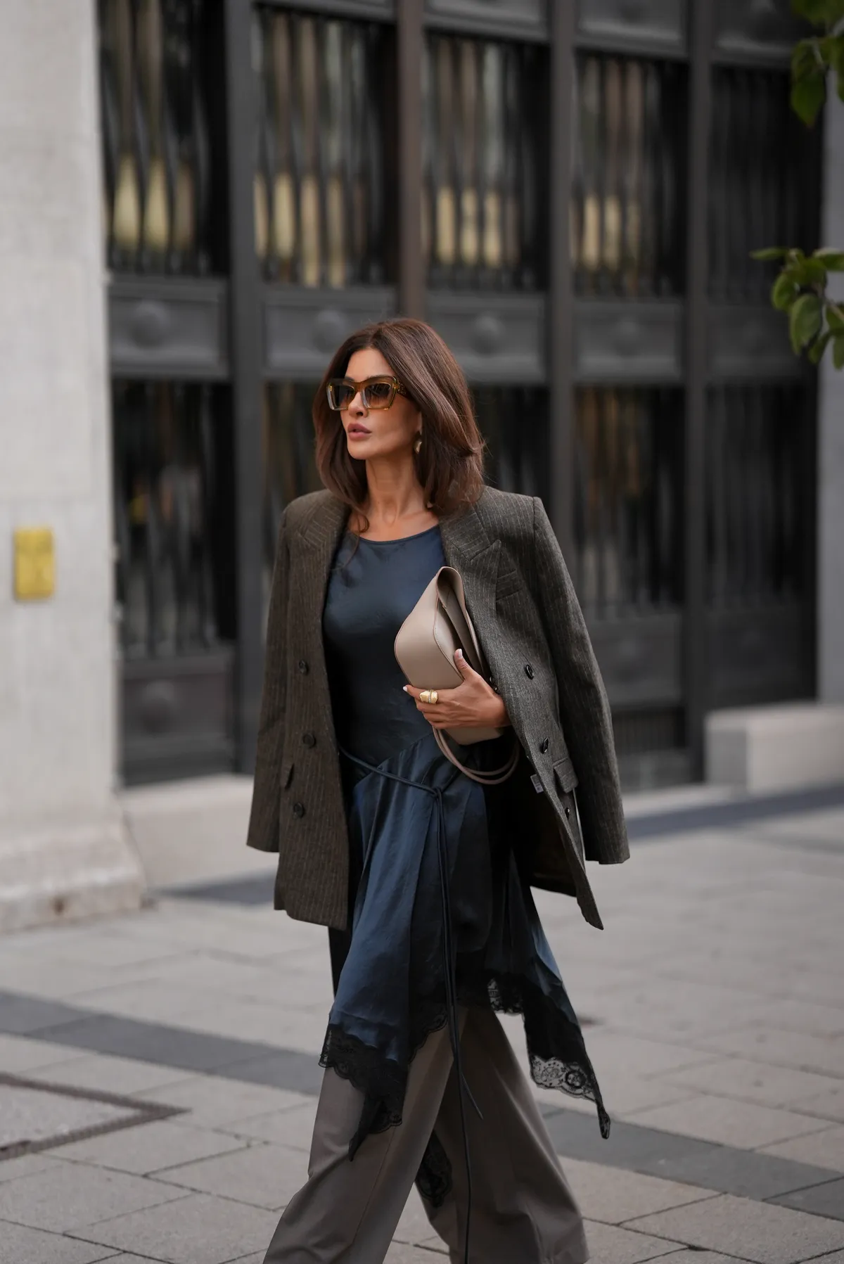 MUNICH, GERMANY - SEPTEMBER 3: Füsun Lindner is seen wearing a dark grey, double-breasted blazer draped casually over the shoulders from Isabel Marant Etoile; a flowing navy silk dress with an asymmetrical handkerchief hem, finished with black lace trim from Acne; grey tailored suit trousers in a straight cut from Acne; a light beige leather handbag with gold hardware and minimal branding from Loewe; oversized brown-tinted sunglasses with chunky frames from Loewe; her brown hair is worn in a straight bob with a center part on September 3, 2025 in Munich, Germany. (Photo by Moritz Scholz/Getty Images)