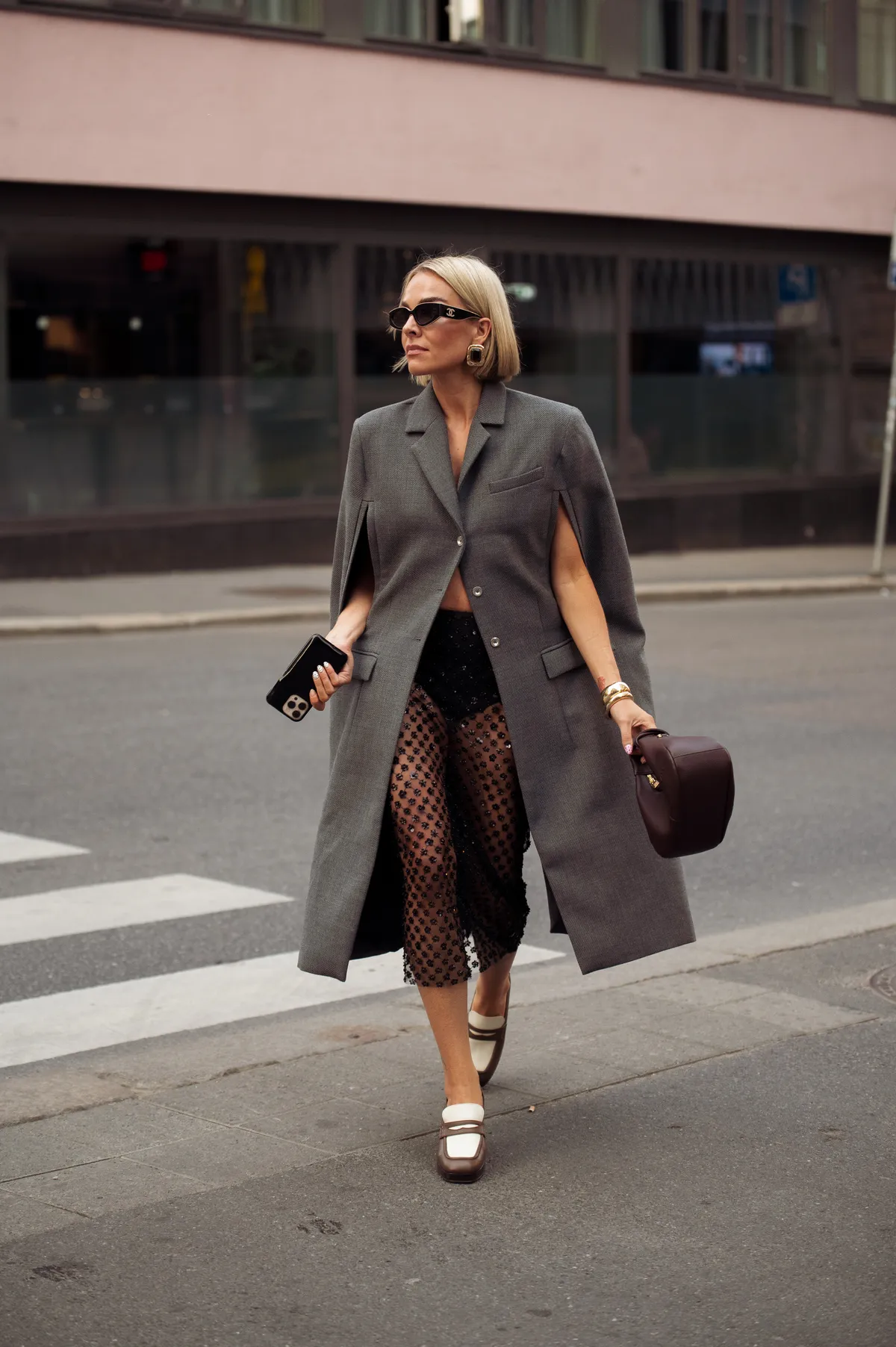 OSLO, NORWAY - AUGUST 26: A guest wears black sheer skirt, brown and white loafers, grey cape coat and brown bag outside Cohme Atelier show during Oslo Fashion Week on August 26, 2025 in Oslo, Norway. (Photo by Raimonda Kulikauskiene/Getty Images)