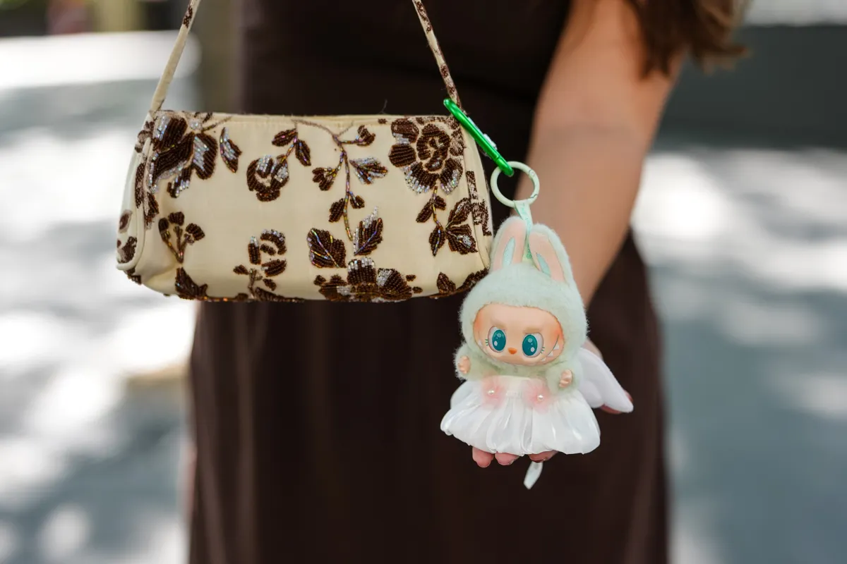 PARIS, FRANCE - JULY 09: A guest wears a strapless brown dress with a fitted bodice and a gathered waistline, creating a flowing silhouette. The hair is styled in loose waves. Sunglasses with dark lenses and a chunky silver necklace are worn. The handbag is beige with intricate brown floral beadwork and features a matching strap. A plush Labubu with a mint green hood and pink inner ears is attached, showcasing a white skirt with pink floral accents, outside Robert Wun, during Paris Fashion Week - Haute Couture Fall/Winter 2025/2026, on July 09, 2025 in Paris, France (Photo by Edward Berthelot/Getty Images)