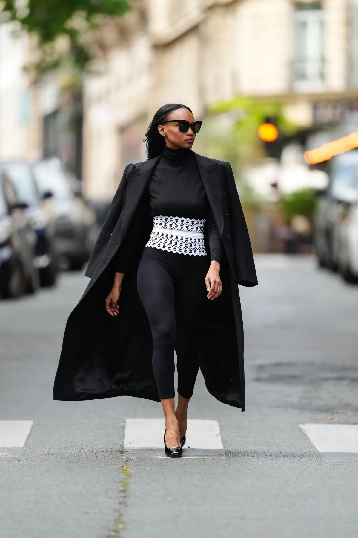 PARIS, FRANCE - MAY 20: Emilie Joseph wears sunglasses, earrings, pink lipstick, a black turtleneck unitard / yoga pants / leggings, a white Alaia printed corset / bustier belt with scalloped edges, a sharply tailored black double breasted long / extra long light wool coat with padded shoulders by COS, maxine buckle embellished glossy patent leather slingback / stiletto pumps / sandals / shoes by Saint Laurent YSL, during a street style fashion photo session, on May 20, 2024 in Paris, France. (Photo by Edward Berthelot/Getty Images)