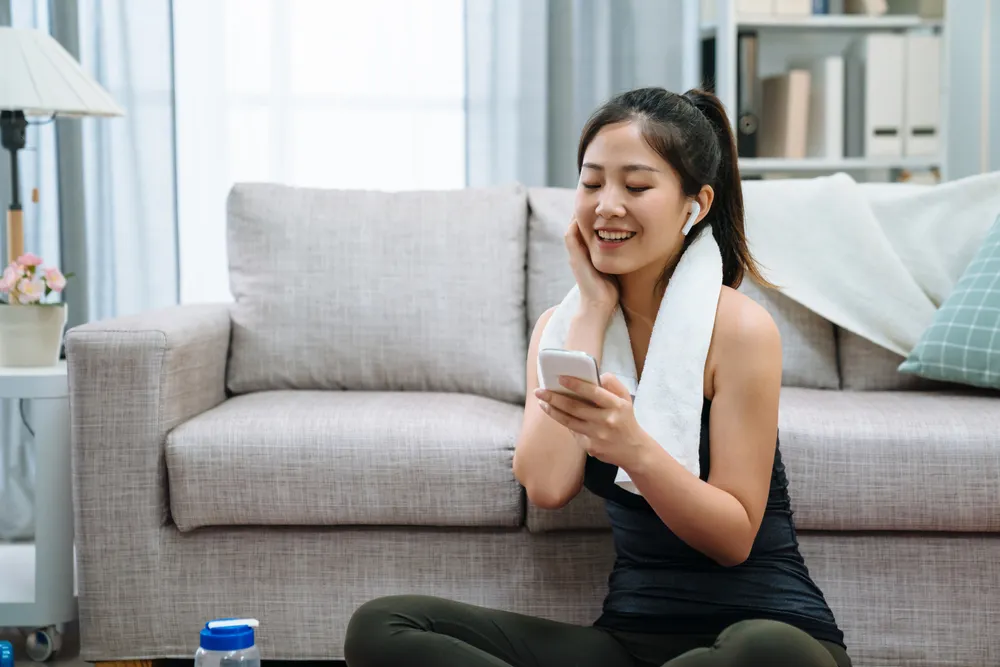 fitness sport training technology lifestyle concept. smiling woman with smartphone and bluetooth earphones enjoy music with towel on neck. joyful lady listen song in wireless headphones on yoga mat