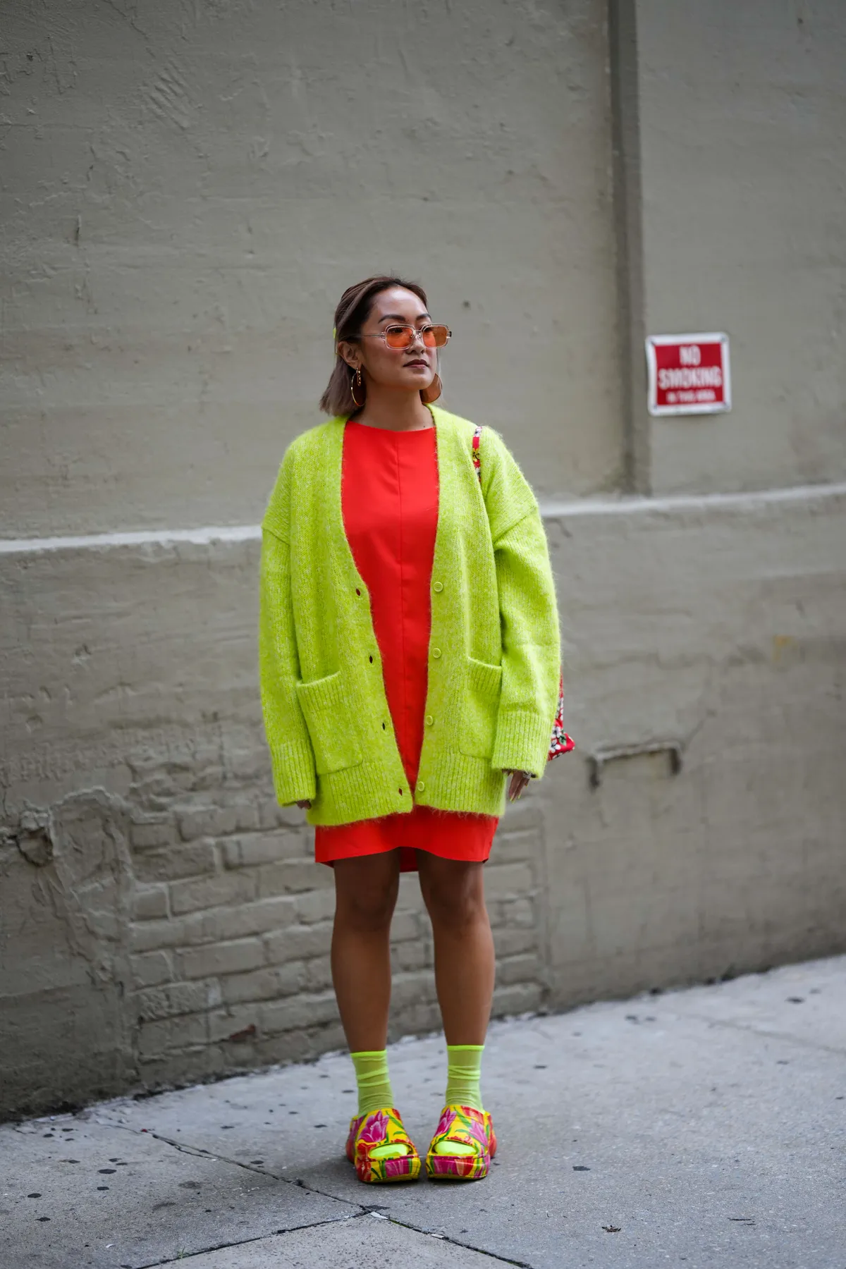 NEW YORK, NEW YORK - SEPTEMBER 13: A guest wears orange sunglasses, gold pendant Fendi earrings, gold earrings, a neon orange short dress, a neon yellow oversized wool cardigan, neon yellow socks, a red with white flower print pattern shoulder bag, yellow with red and pink flower painted pattern open toe cap / plastic mules , outside COS, during New York Fashion Week, on September 13, 2022 in New York City. (Photo by Edward Berthelot/Getty Images)