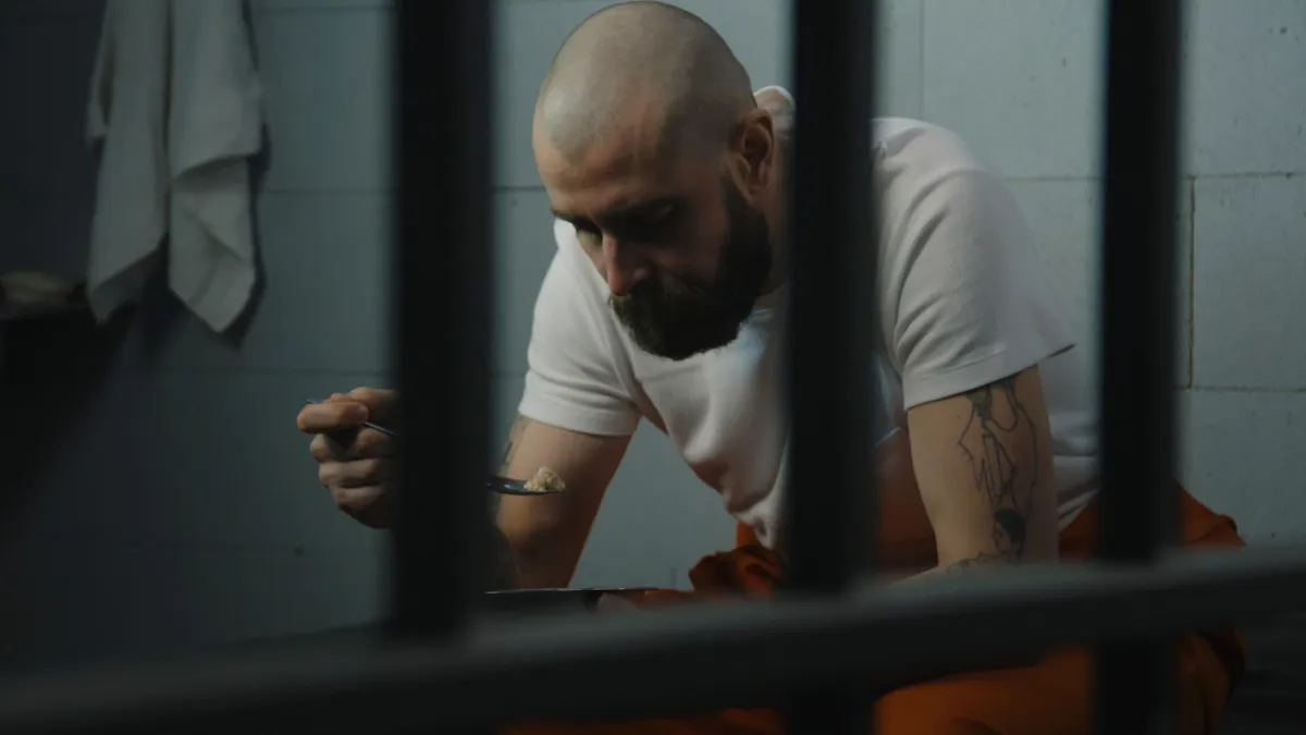 Inmate in tattoos in orange uniform sits on bed in jail cell, eats prison food from iron bowl. Male prisoner serves imprisonment term for crime in prison. Detention center or correctional facility.