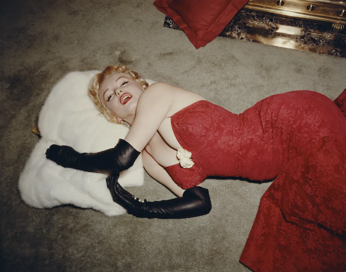 1955: EXCLUSIVE American actor Marilyn Monroe (1926 - 1962), wearing a red brocade evening gown and long black gloves, lying on a carpet and using a white fur stole as a pillow. (Photo by Gene Lester/Getty Images)