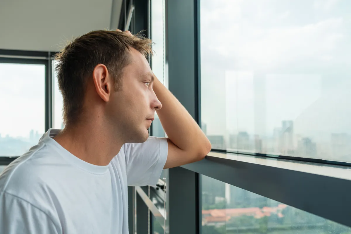 Unhappy frustrated man suffering from depression and bad news looking at window with city view. Tired sad male at home. Mental health, life problems, emotional stress concept