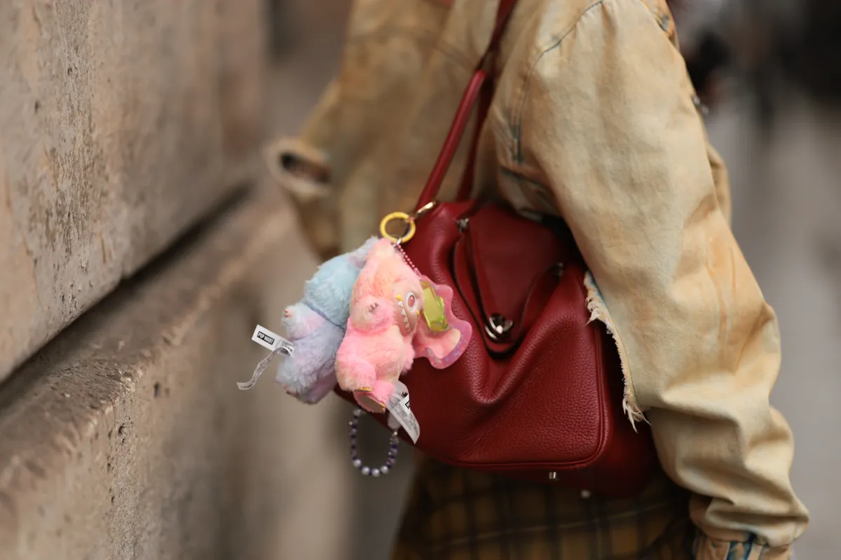 PARIS, FRANCE - JUNE 25: A Fashion Week guest is seen wearing an oversized denim jacket in a blue-brown wash from EgonLab; a rectangular red leather “Lindy” shoulder bag from Hermès featuring two pastel-colored Labubu monster charms; and an ochre-brown checked long shirt underneath during the Jeanne Friot Menswear Spring/Summer 2026 show as part of Paris Fashion Week on June 25, 2025 in Paris, France. (Photo by Jeremy Moeller/Getty Images)