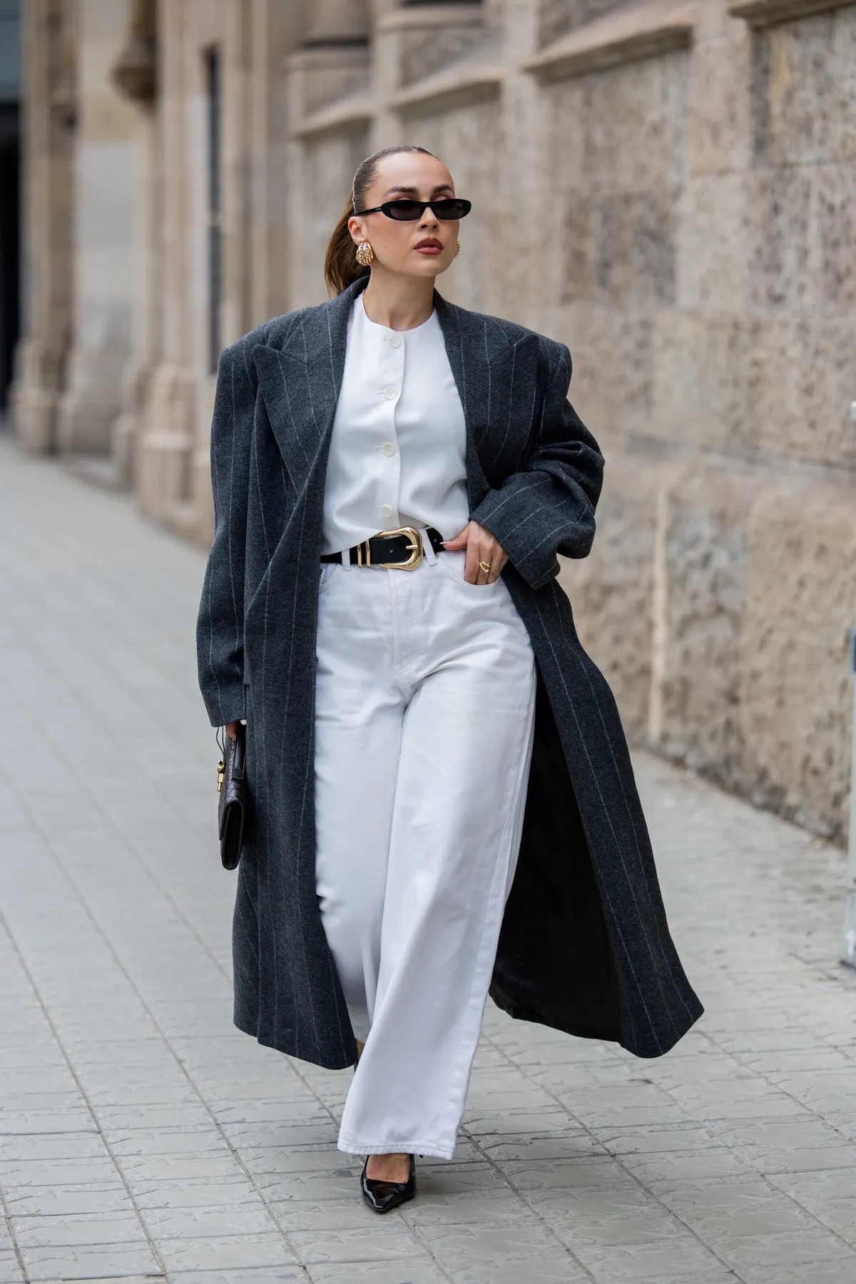 BARCELONA, SPAIN - MARCH 27: Anna Wein wears grey striped oversized coat Bazhan, heeled shoes YSL, bag Bottega Veneta Long Andiamo intrecciato leather clutch, white jeans ARKET, golden earrings, sunglasses, belt on March 27, 2025 in Barcelona, Spain. (Photo by Christian Vierig/Getty Images)