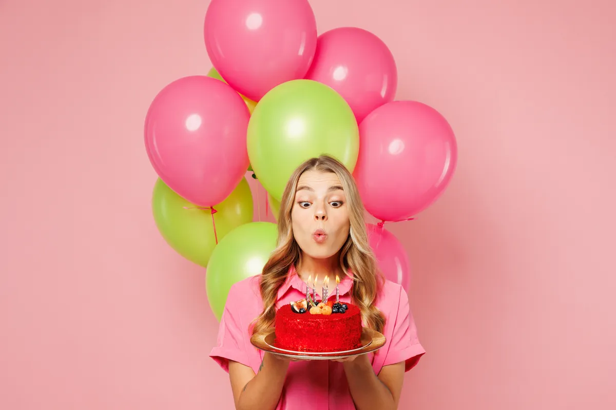 Young woman wears shirt celebrating birthday holiday and hold bunch of colorful air balloons, red cake blow out candles making wish isolated on plain pastel light pink background. Lifestyle concept