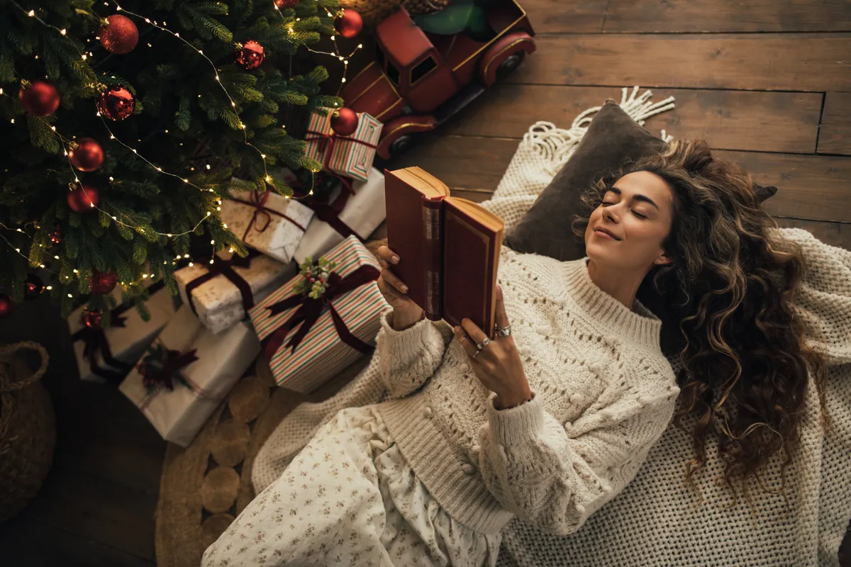 Young beautiful woman reading book, lying near Christmas tree in a cozy decorated room.