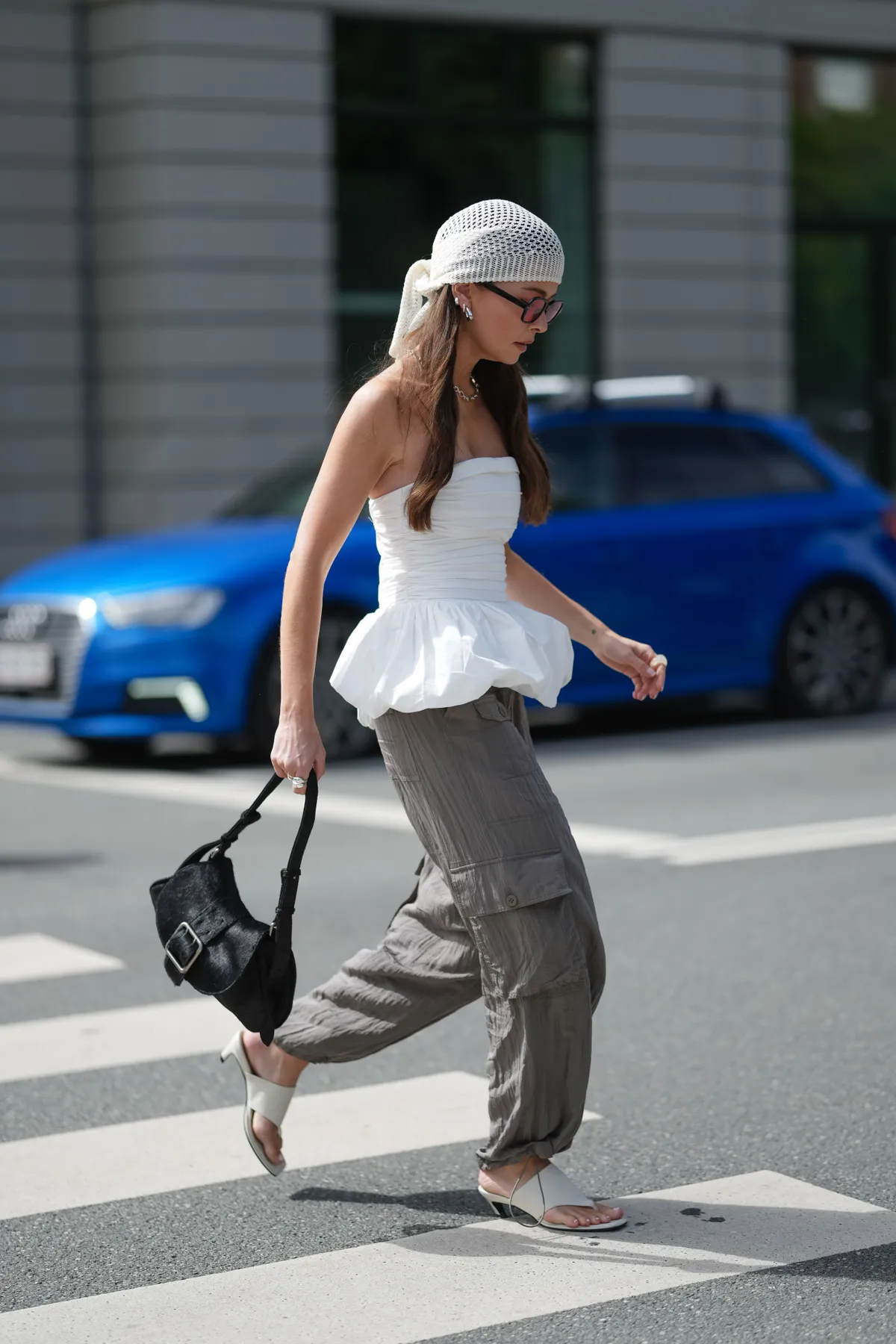 COPENHAGEN, DENMARK - AUGUST 06: A guest wears a white, textured headscarf tied at the back. Hair is styled long and loose. Sunglasses feature a black frame. Earrings are small and silver-toned. A strapless white top with a ruched bodice and peplum hem is worn. Loose-fitting cargo pants in a taupe shade with large side pockets are paired. A black handbag with a visible buckle is carried. Footwear consists of white open-toe mules with a low heel, outside Munthe, during Copenhagen Fashion Week, on August 06, 2025 in Copenhagen, Denmark (Photo by Edward Berthelot/Getty Images)