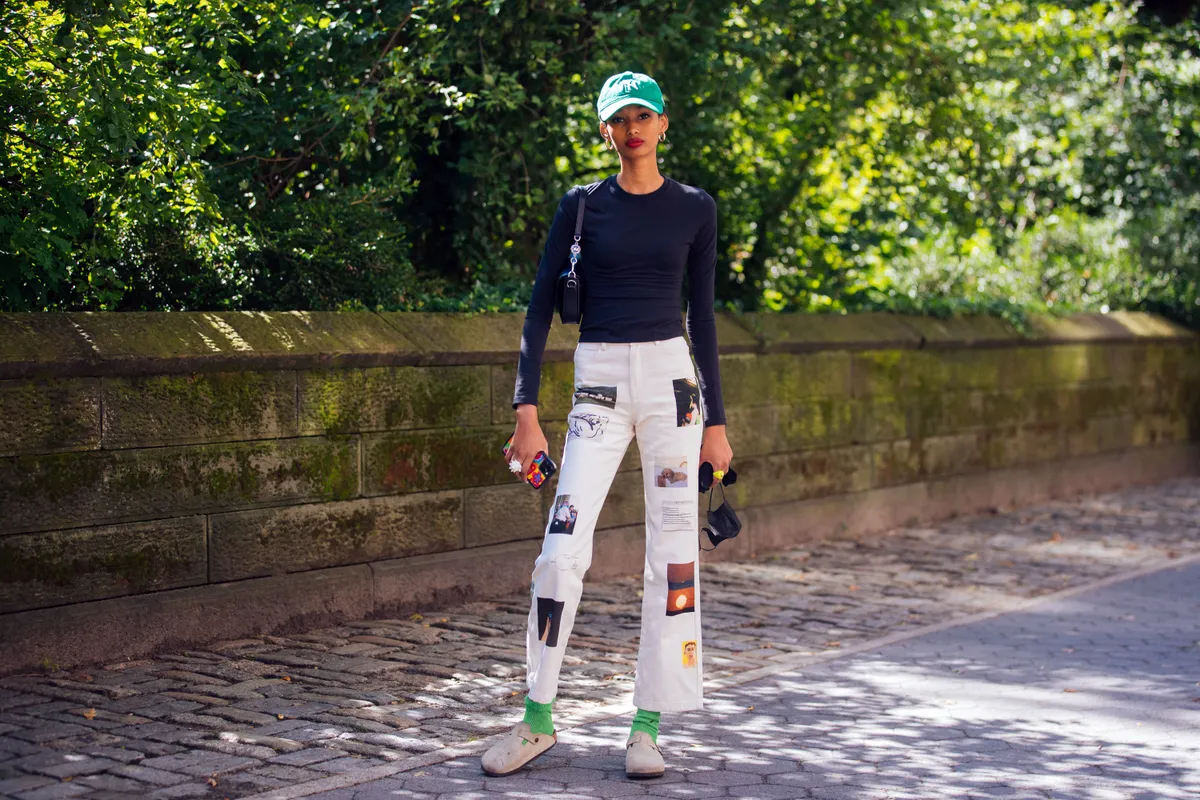 NEW YORK, NEW YORK - SEPTEMBER 10: Model Ugbad Abdi wears a green Yankees cap, gold earrings, black fitted long-sleeve top, black Michael Kors shoulder bag, white Paloma Wool high waist straight Friendship pants with art and photos by Alejandra Smits, Carlota Guerrero, Camila Falquez, Leticia Sala, María Gaminde, Tana Latorr, María Sosa, Olga De La Iglesia, and Paloma Lanna, green socks, and tan suede clogs after the Michael Kors show on September 10, 2021 in New York City. (Photo by Melodie Jeng/Getty Images)