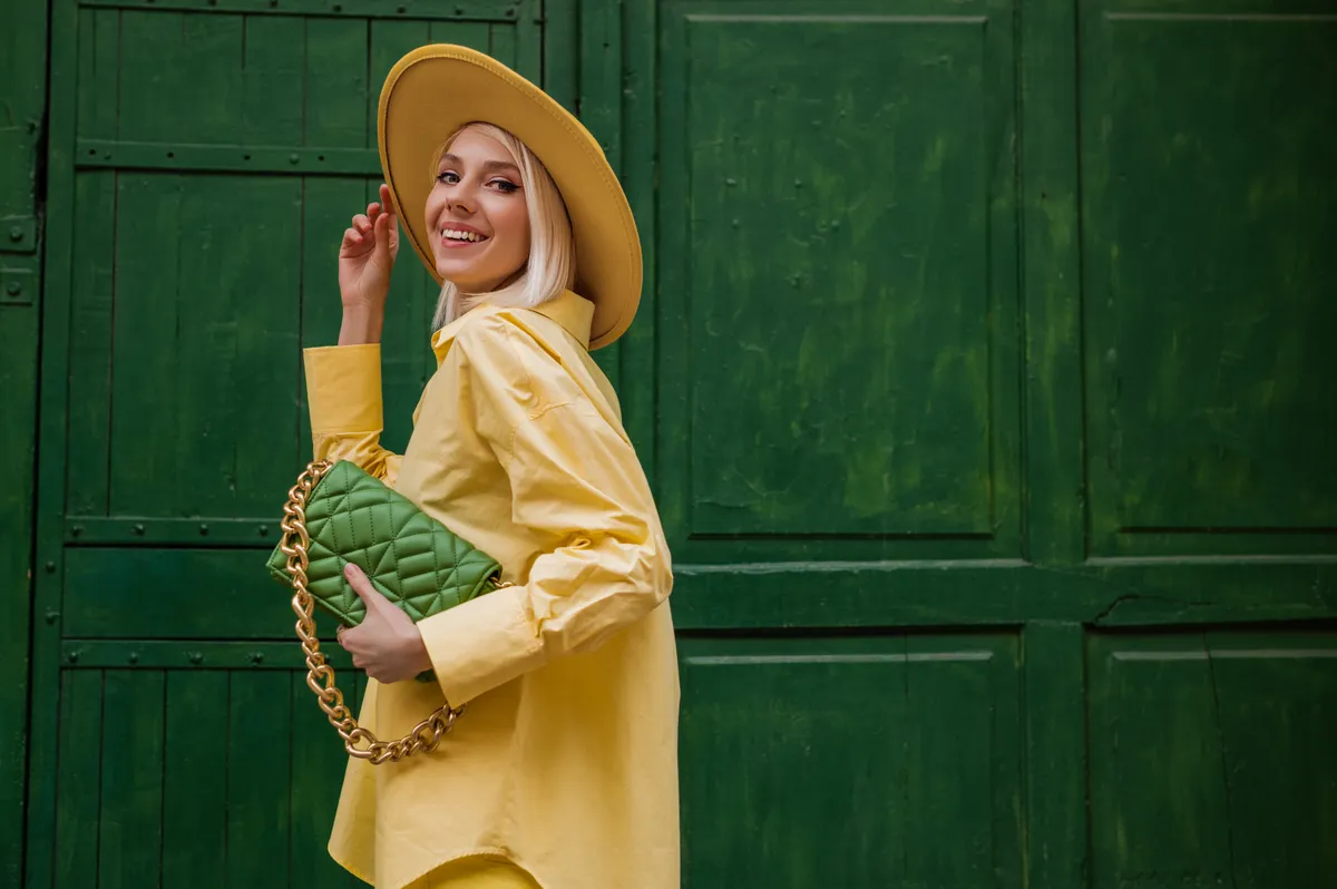 Joyful smiling woman wearing yellow hat, classic shirt, holding quilted faux leather green bag with chain, posing on green background in street. Copy, empty space for text