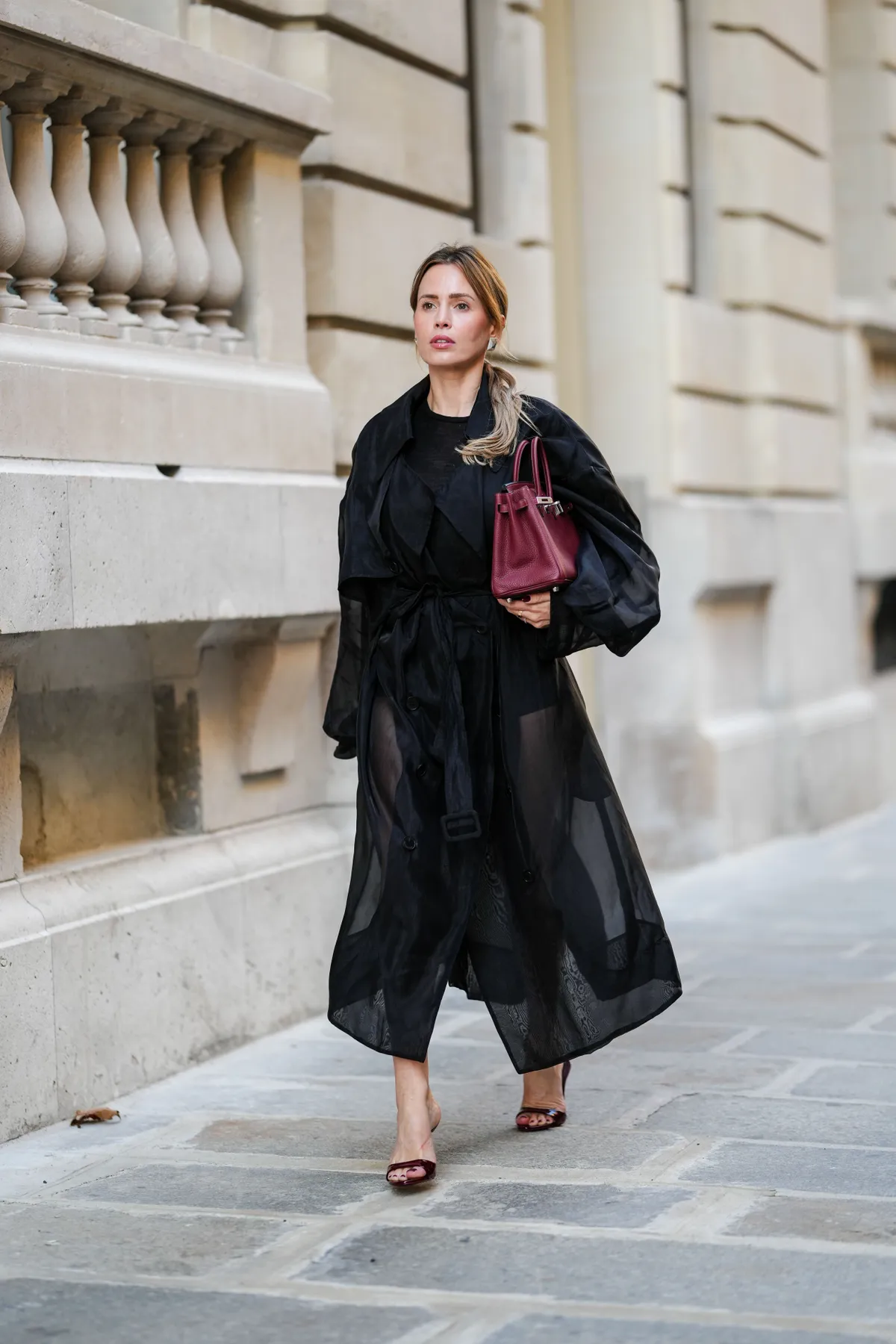 PARIS, FRANCE - OCTOBER 23: Virginie Conte wears a black transparent the Frankie shop trench coat, silver Bruna The Label earrings, a black COS top, black The Frankie Shop shorts, a burgundy leather Birkin Hermes bag, burgundy leather Paris Texas open toes high heels, during a street style fashion photo session, on October 23, 2024 in Paris, France. (Photo by Edward Berthelot/Getty Images)